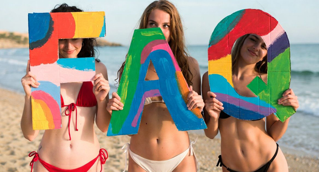 Tre studentesse sulla spiaggia tengono lettere colorate FAQ durante un soggiorno linguistico LISA! a Malta