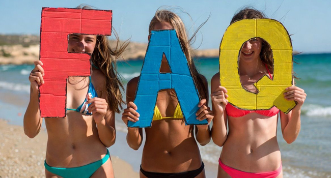 Soggiorno linguistico per studenti a Malta: FAQ per St. Julian’s Tre studentesse di un soggiorno linguistico per studenti a Malta sulla spiaggia di St. Julian’s con grandi lettere FAQ