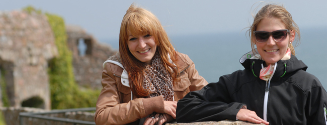 Zwei Frauen lächseln vor dem Meer und alten bewachsenen Steinen in die Kamera bei einer Sprachreise Jersey