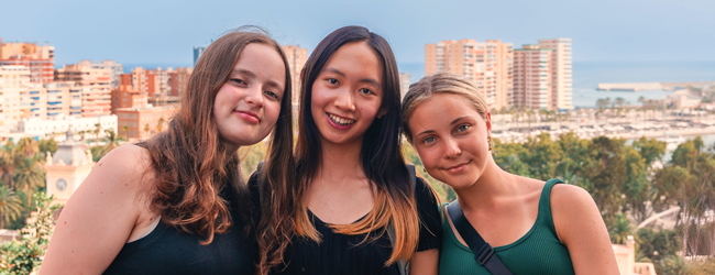 In der Stadt unterwegs bei einer LISA! Schülersprachreise in Barcelona Drei Mädchen lächeln in die Kamera vor einer städtischen Kulisse im Hintergrund das Meer in Barcelona